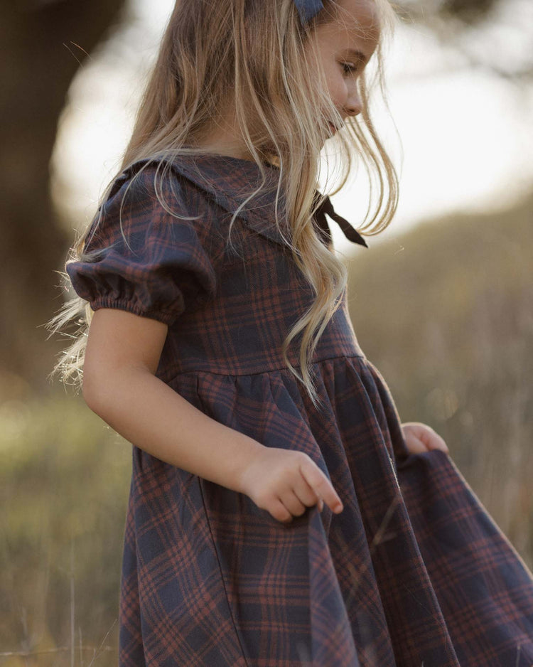 A young girl with long blonde hair stands outdoors in soft sunlight, looking down as she holds the edge of her *camille dress in midnight plaid* by rylee + cru—crafted from 100% cotton with puffed sleeves and a delicate bow.