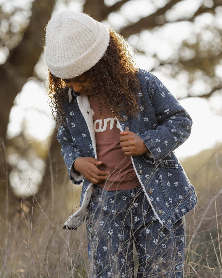 A young child with curly hair stands outdoors in tall grass, looking down while wearing the rylee + cru long chore coat in heart ditsy—a white knit hat and matching pants complete the look.