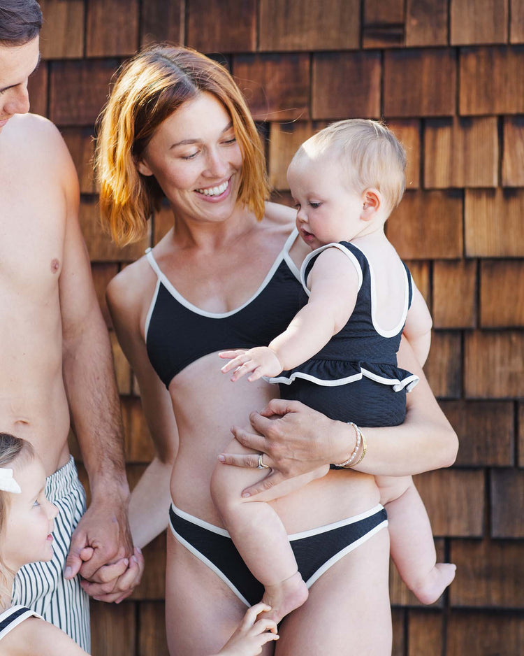 A woman in a rylee + cru skirted one-piece in black smiles while holding a baby in a matching swimsuit, with an adult and child partially visible beside her against a wooden shingle wall.
