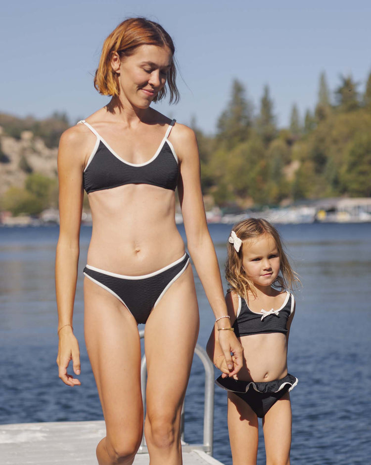 A woman and a young girl, hand in hand on a lakeside dock, wear matching rylee + cru daisy bikinis in black made from recycled polyamide, with sunny trees and hills in the background.