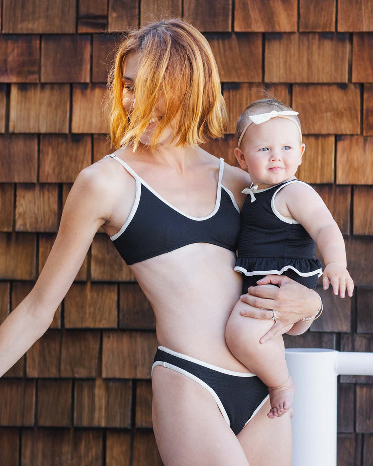 A woman wears the rylee + cru skirted one-piece in black while holding a baby in a matching UPF 50+ swimsuit and headband. They stand before a wall with wooden shingles, the woman’s face partially turned away.