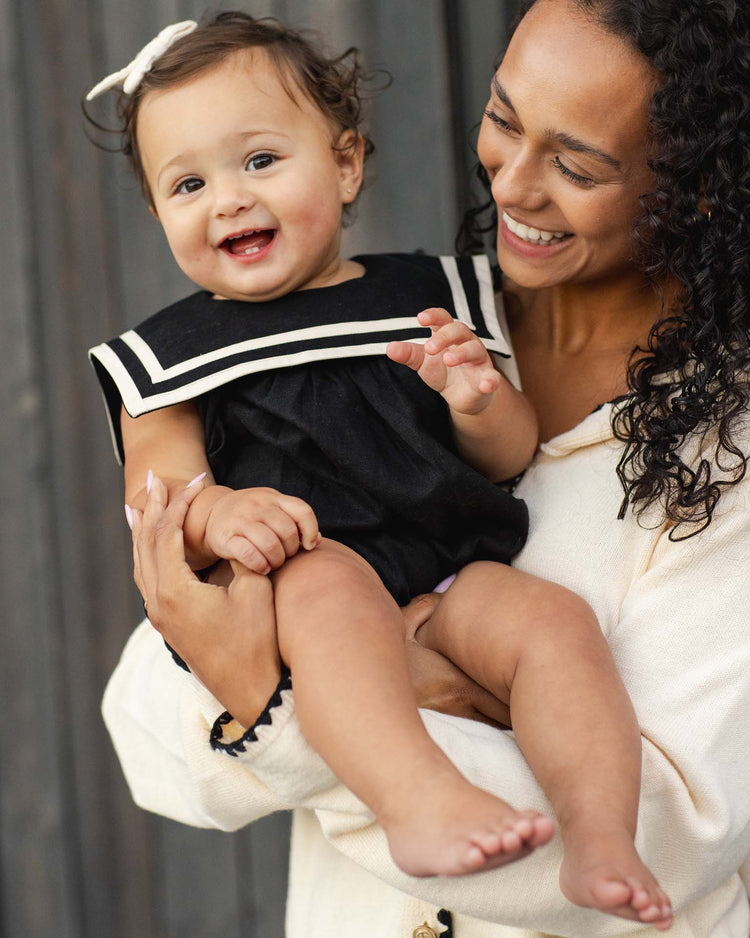 A smiling woman with curly hair holds a happy baby girl wearing the rylee + cru sailor romper in black, featuring white trim and a white bow. Both appear joyful against a dark wooden background.
