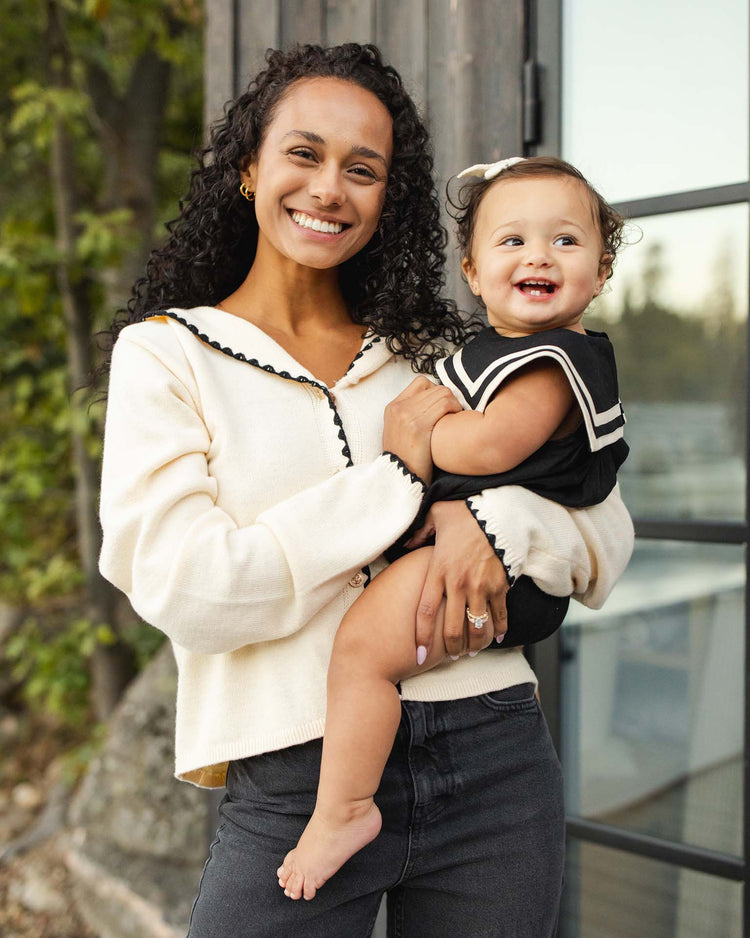 A smiling woman with curly hair holds a happy baby girl wearing the rylee + cru sailor romper in black and a white bow. The woman wears a cream sweater and dark jeans. They stand outside near a building with glass windows.