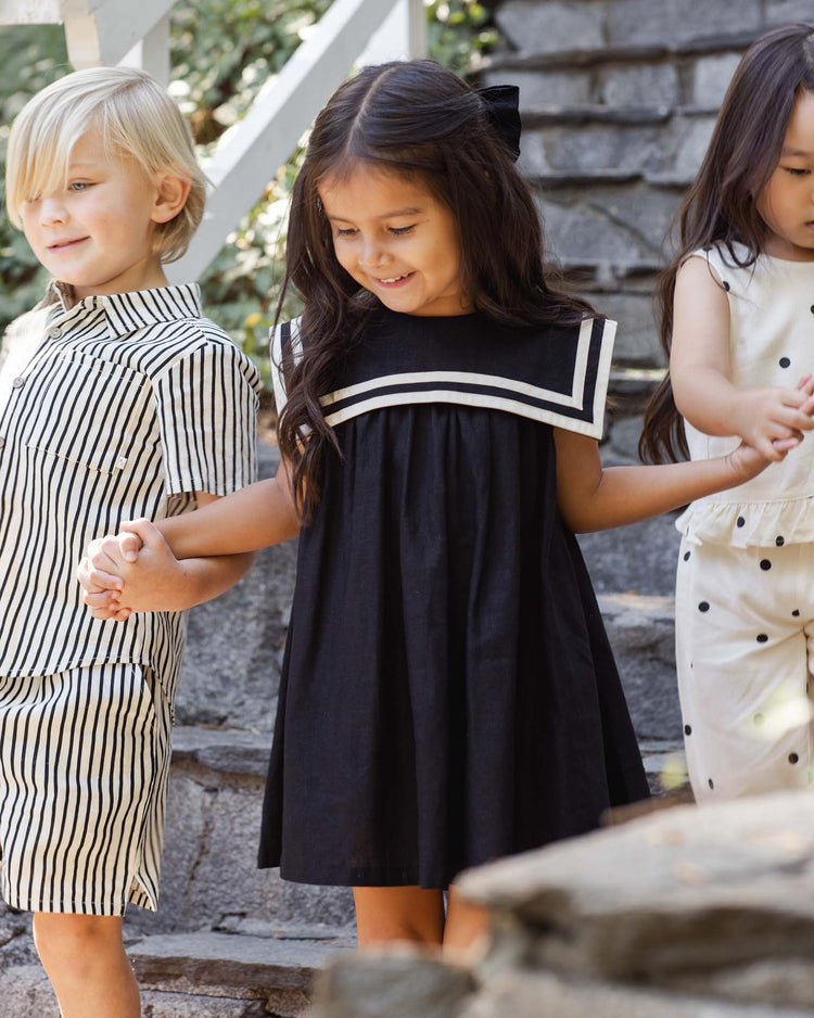 Three young children holding hands walk down outdoor stone steps. The girl in the middle wears the rylee + cru sailor dress in black, while the others wear playful striped and dotted outfits.
