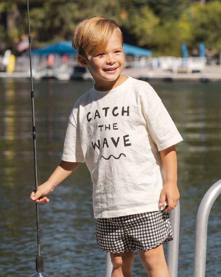A blonde-haired child smiles by the water, holding a fishing rod and wearing rylee + cru’s relaxed tee in catch the wave with checkered shorts. Trees and boats are visible in the background.