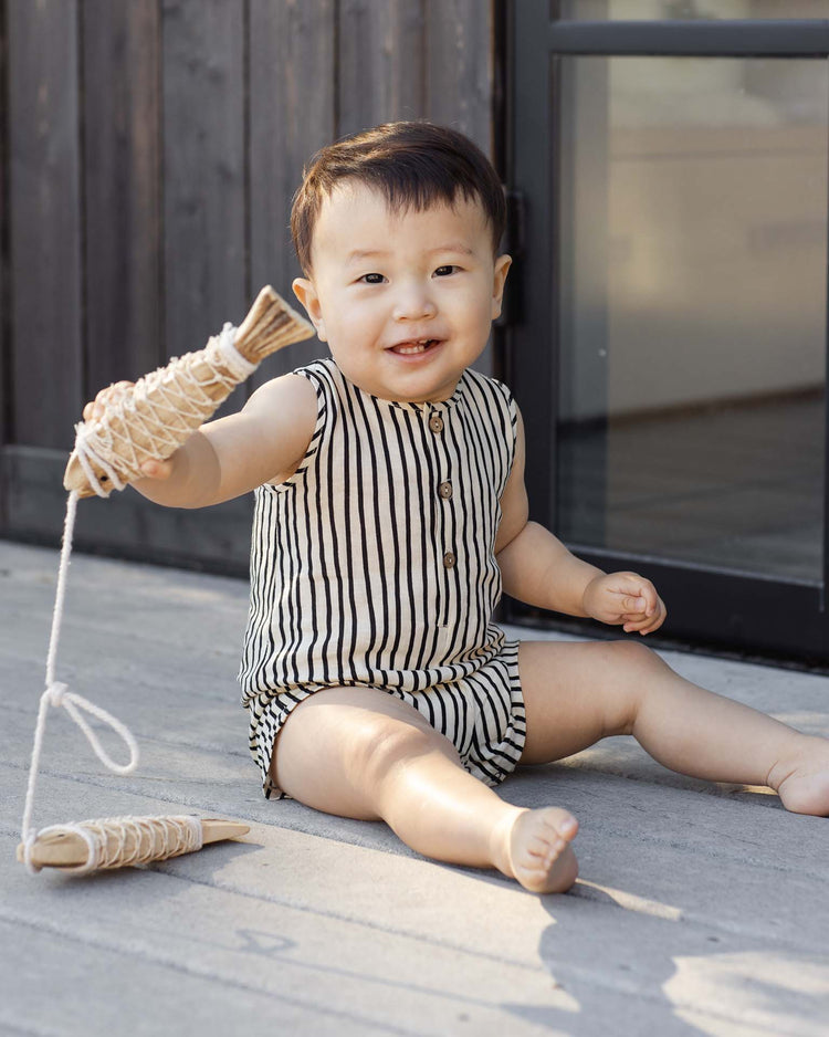 A smiling toddler wearing the rylee + cru mavrick romper in wavy stripe sits on a wooden deck holding a toy fish, with a modern door and wooden wall in the background.