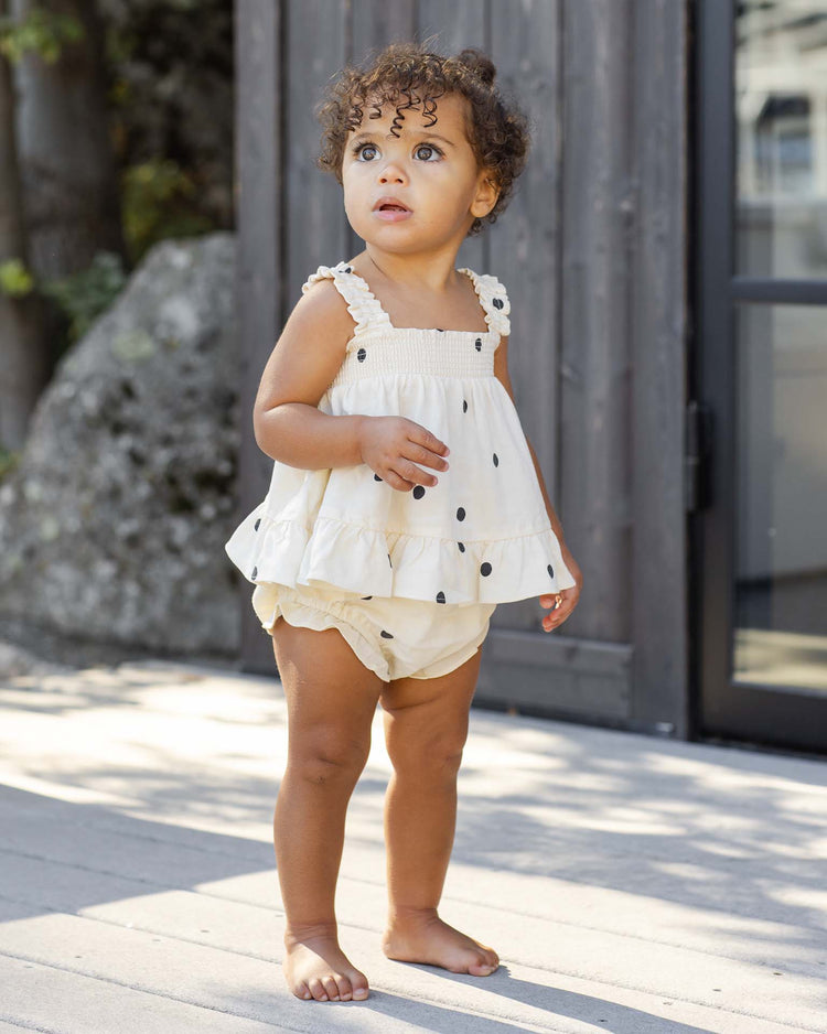 A young child with curly hair stands barefoot on a wooden deck, wearing the rylee + cru lala set in polka dot with flutter bloomers and subtle ruffle detail, gazing upwards with curiosity. A wooden wall and rocks form the background.
