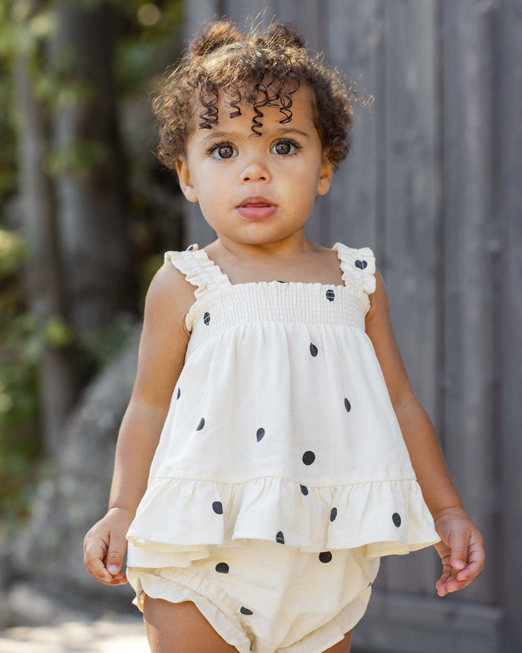 A young child with curly hair and light eyes stands outdoors in a rylee + cru lala set in polka dot, featuring a white top with ruffle details and matching flutter bloomers, looking directly at the camera.