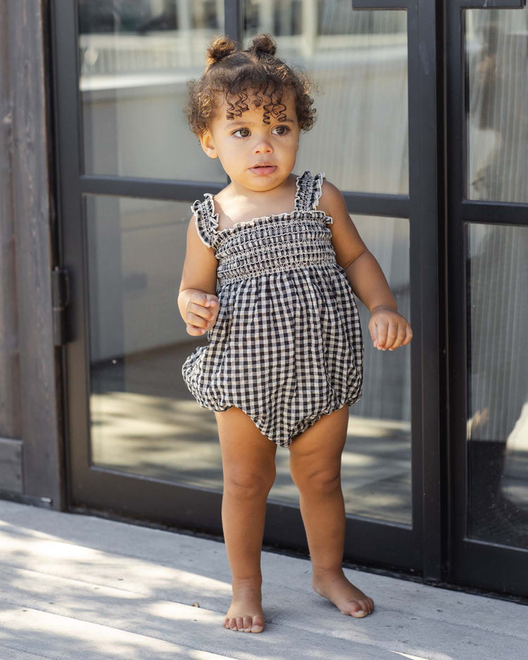 A toddler with curly hair in two buns stands barefoot on a patio in front of a glass door, wearing the rylee + cru julia romper in black check.