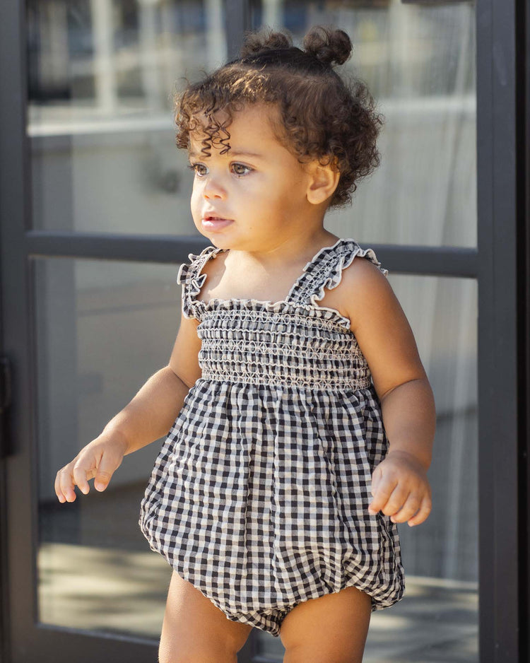 A toddler with curly hair wears the rylee + cru julia romper in black check, standing by a glass door and gazing thoughtfully to the side.