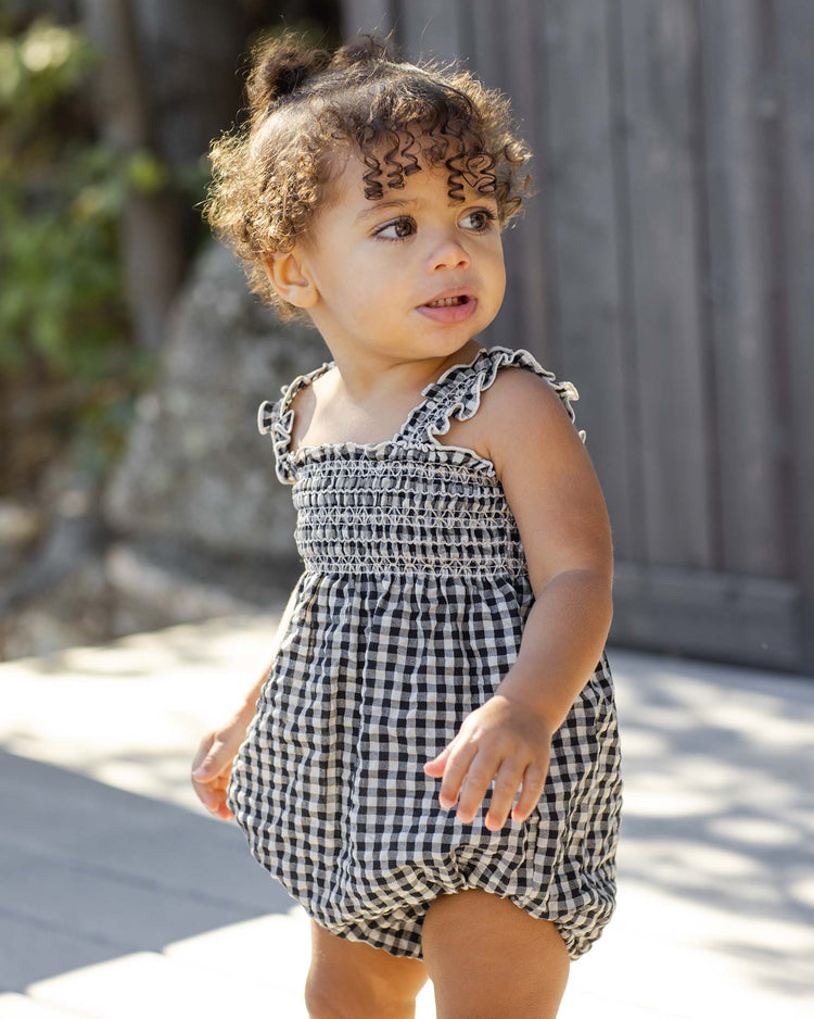 A young child with curly hair stands outdoors on a sunny day, wearing the rylee + cru julia romper in black check and looking to the side with a slight smile.