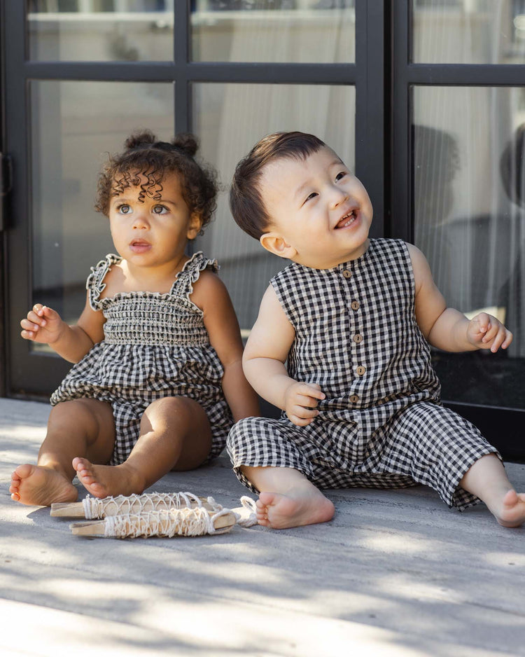 Two young toddlers sit on a porch before glass doors. One, barefoot and smiling with curly hair, wears the rylee + cru button jumpsuit in black check; the other, with straight hair, also sports this matching one-piece.