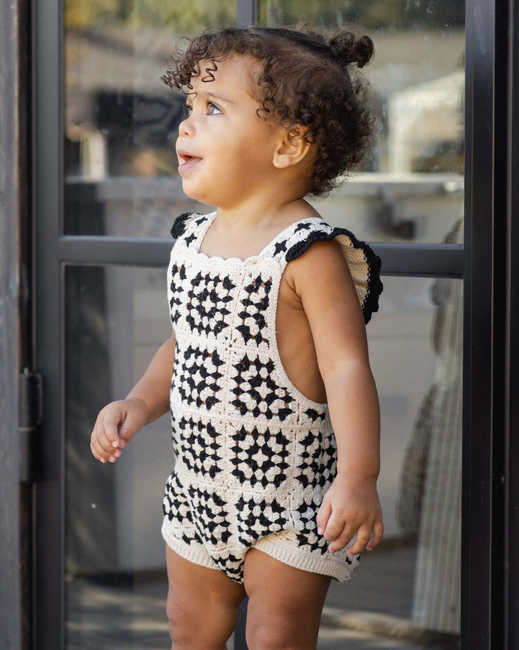 A young child with curly hair stands in front of a glass door, wearing the rylee + cru crochet romper in abstract and gazing up with a curious expression and timeless vintage charm.