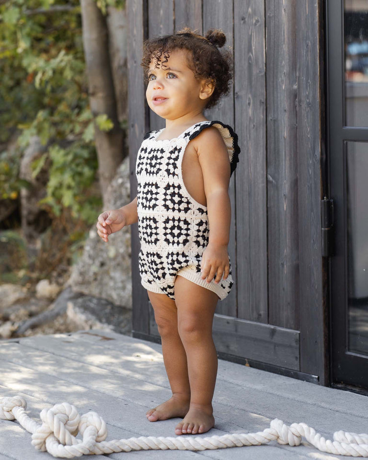 A young child stands barefoot on a patio near greenery and a wooden structure, wearing the rylee + cru crochet romper in abstract, made from 100% cotton with vintage charm. A thick rope lies nearby.