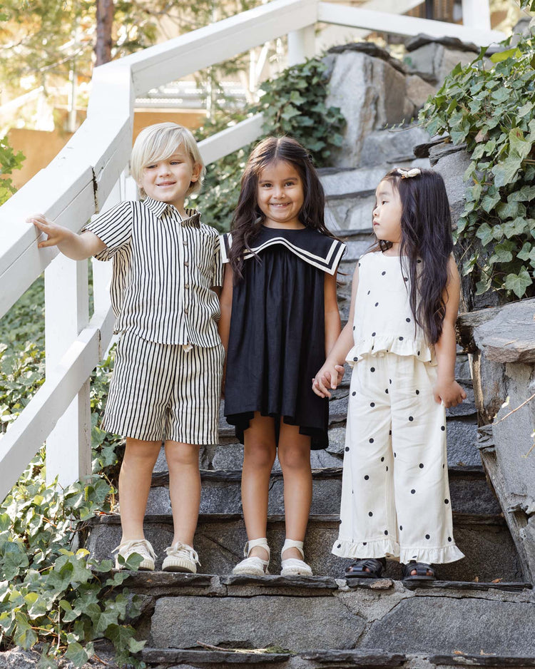 Three children stand on stone steps, smiling. The boy wears the rylee + cru lapel collared shirt in black check, while one girl is in a black dress and the other in a white polka-dot outfit.