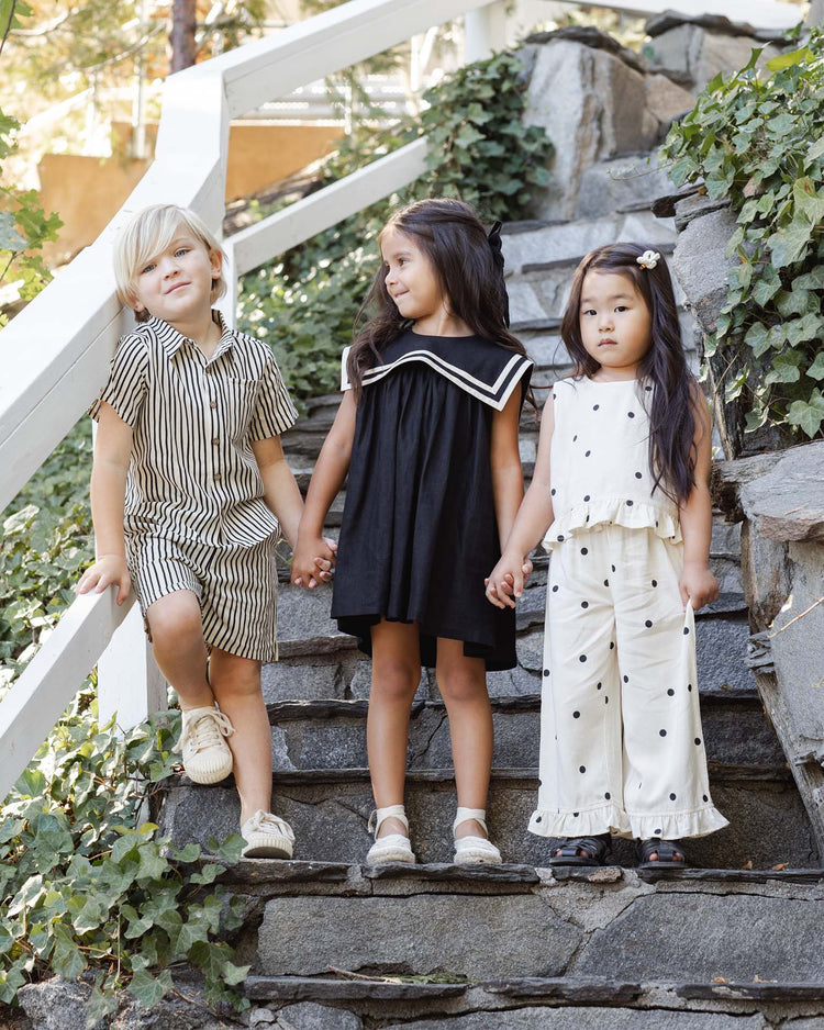 Three young children stand on outdoor stone steps with green ivy. One wears the rylee + cru oceanside top in polka dot, one a sleeveless ruffle cotton top, and one a black dress with a sailor collar. They hold hands and smile.