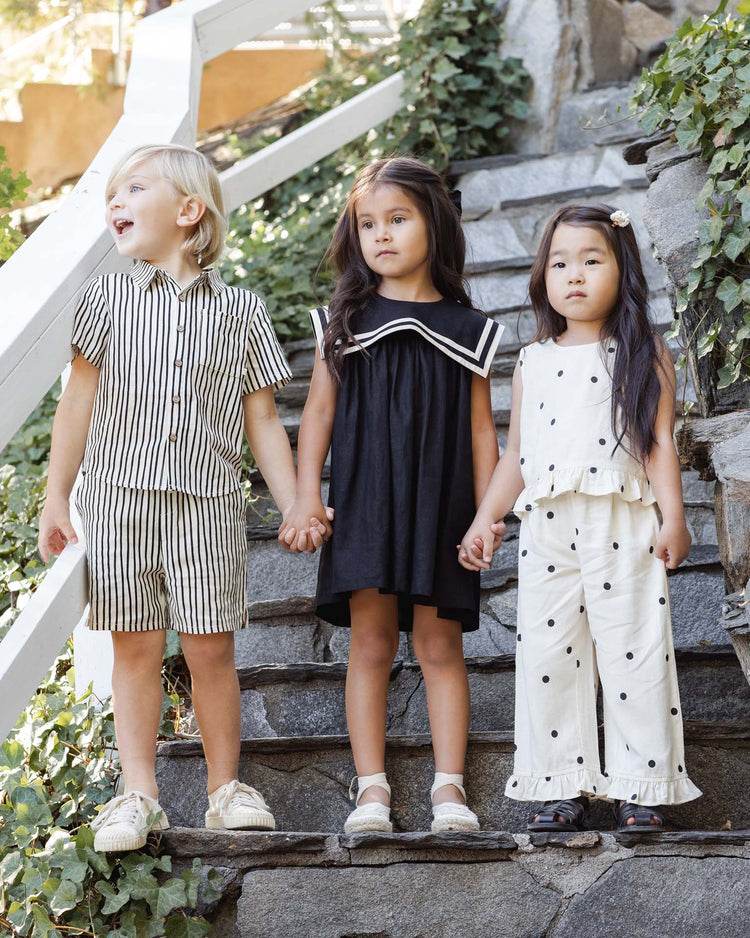 Three young children stand on stone steps holding hands. The girl in the middle wears the rylee + cru sailor dress in black, while the others sport playful outfits. Lush green vines surround them, creating a charming scene.