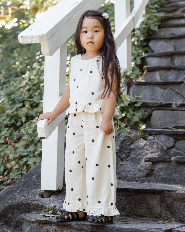 A young girl stands on stone steps surrounded by greenery, wearing rylee + cru’s wide leg pant in polka dot and black sandals, paired with a matching cream-colored top.