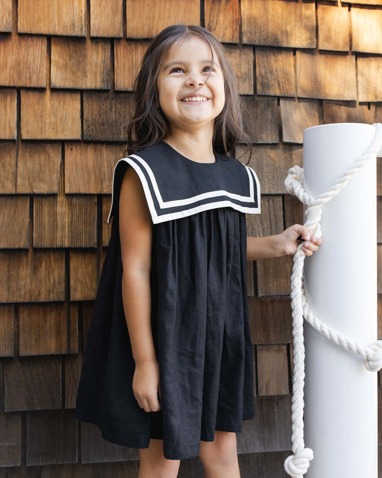 A young girl smiles in front of a shingled wall, wearing the rylee + cru sailor dress in black with a white collar, and holding a white post wrapped in thick rope.