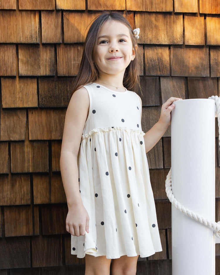 A young girl with long brown hair smiles beside a white post, wearing the rylee + cru harper dress in polka dot with coordinating bloomers, standing in front of a wooden shingle wall.