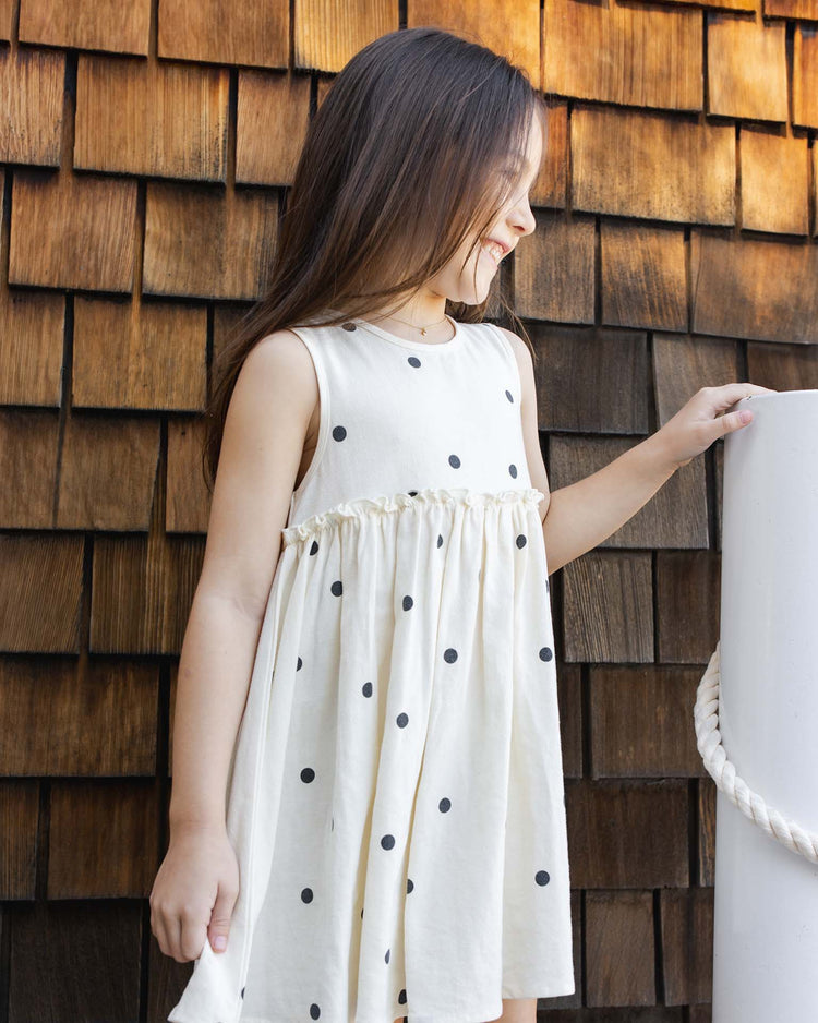 A young girl with long brown hair smiles and looks to the side while wearing the rylee + cru harper dress in polka dot, standing by a wooden shingle wall and holding a white post with rope.