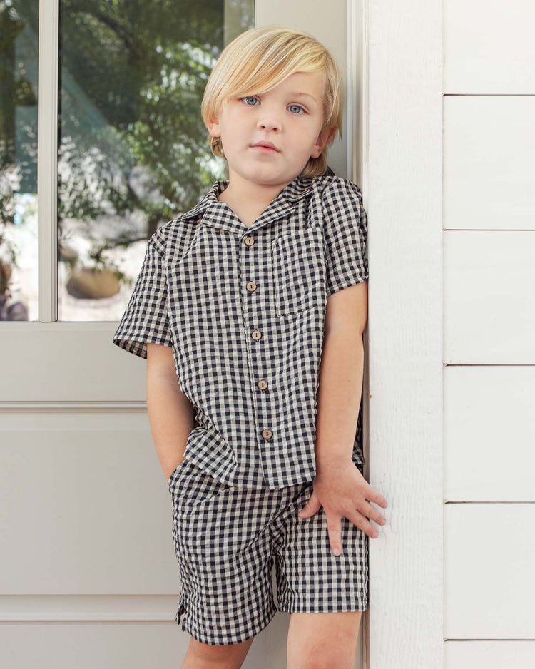 A young child with blond hair stands by a white wall and door, wearing the rylee + cru lapel collared shirt in black check—short-sleeved and button-up—with matching shorts, hands in pockets and a neutral expression.