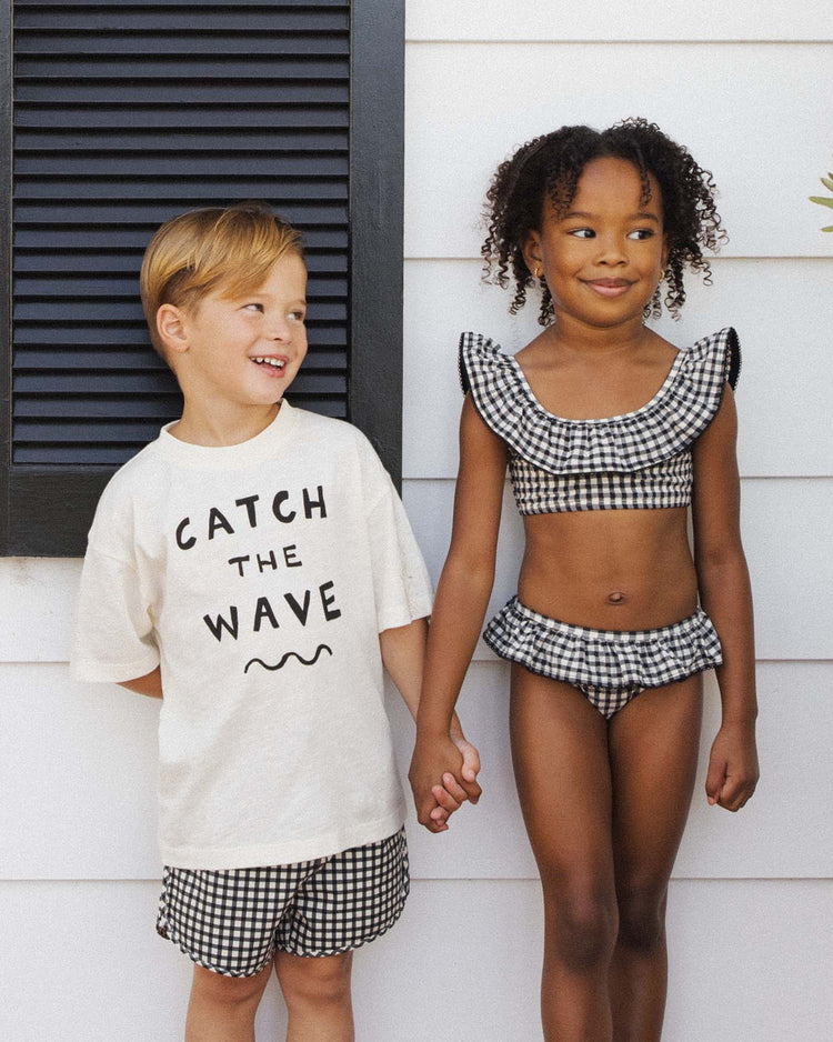 A boy and girl stand holding hands by a white wall. The boy wears the rylee + cru relaxed tee in catch the wave with checkered shorts, while the girl sports a checkered ruffle bikini. Both look cheerful and summery.