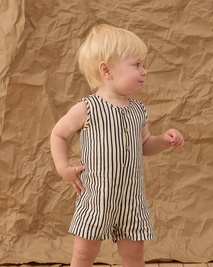 A young child with blonde hair stands sideways before a crumpled brown paper backdrop, wearing the rylee + cru mavrick romper in wavy stripe—a slim cut, sleeveless outfit with vertical stripes and wood-colored buttons.