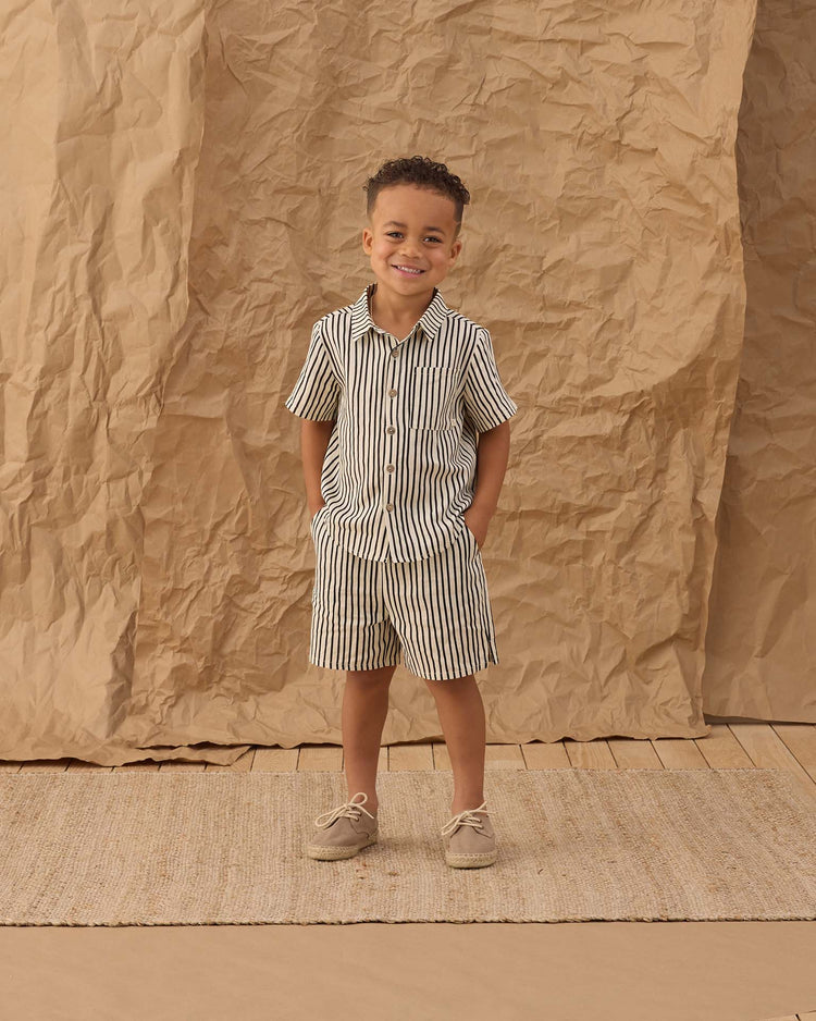 A young boy smiles with his hands in his pockets, wearing the rylee + cru collared short sleeve shirt in wavy stripe and shorts, beige shoes, standing on a woven rug before a crumpled brown paper backdrop.
