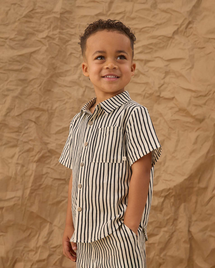 A young boy smiles while looking upward, wearing the rylee + cru collared short sleeve shirt in wavy stripe and matching shorts, standing before a crumpled brown paper background.