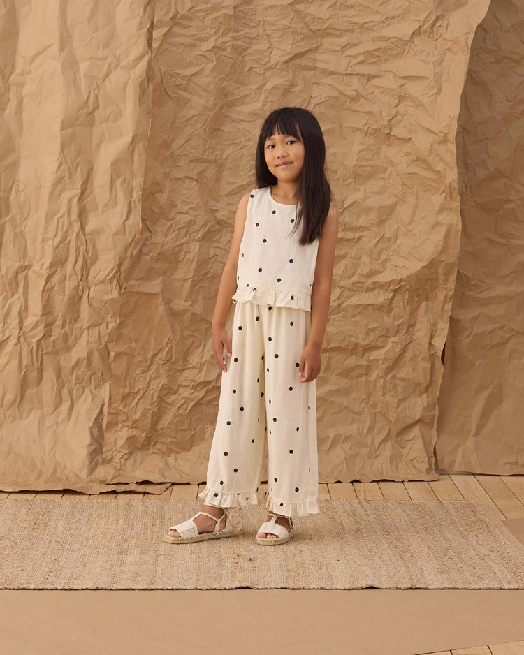 A young girl stands on a woven rug in rylee + cru’s wide leg pant in polka dot, paired with a white sleeveless top. A crumpled brown paper backdrop rests behind her on the wooden floor.
