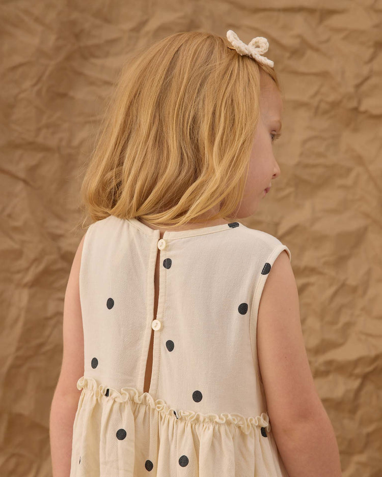A young girl with blonde hair and a cream bow, seen from behind, wears the rylee + cru harper dress in polka dot. She stands against a backdrop of crumpled brown fabric.