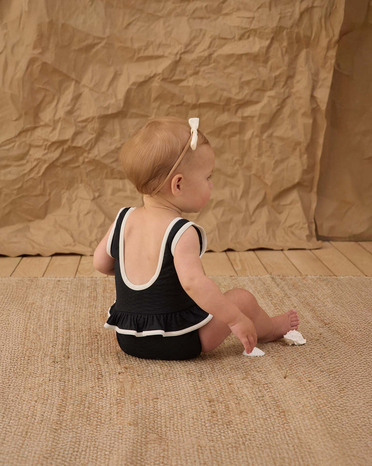 A baby in a rylee + cru skirted one-piece in black with a bow headband sits on a woven rug, facing away from the camera against a crumpled brown paper backdrop.