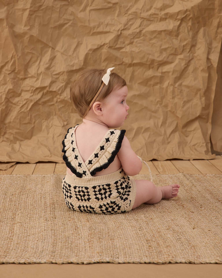 A baby with light brown hair sits on a woven mat, facing away, wearing the rylee + cru crochet romper in abstract and a cream headband. The vintage mood is set by a crumpled brown paper background.