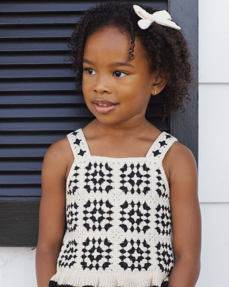 A young girl with curly hair and a cream bow stands in front of a dark shutter, wearing the rylee + cru kira set in abstract—a sleeveless crochet knit top with black and cream squares, paired with cotton shorts—as she smiles slightly, looking to the side.