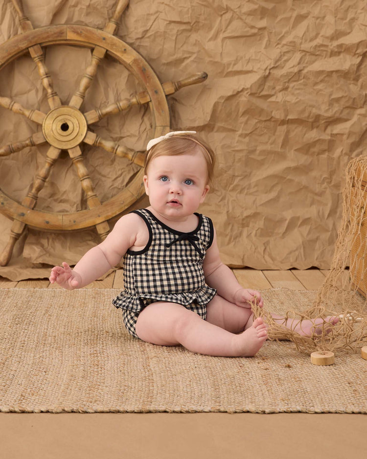 A baby in the rylee + cru skirted one-piece in black gingham sits on a woven mat, with a ship’s wheel and fishing net behind them, against a crumpled brown paper backdrop.