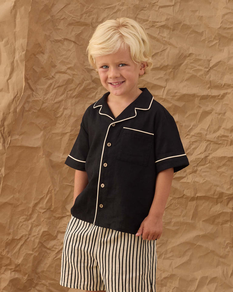 Young child with blond hair wears the rylee + cru lapel collared shirt in black, smiling as they stand before a crumpled brown paper backdrop.