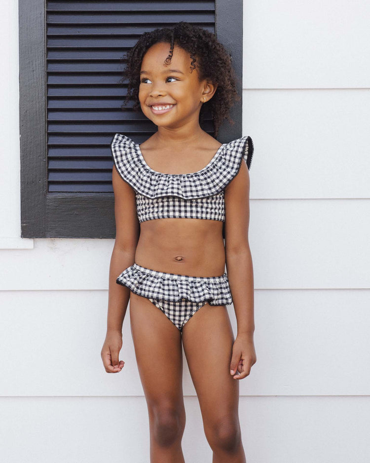 A young girl smiles in front of a white wall, wearing the rylee + cru margot bikini in black gingham.