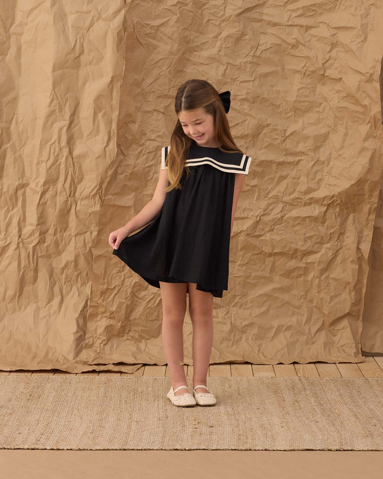 A young girl stands on a woven rug against a brown paper backdrop, wearing the rylee + cru sailor dress in black with a white collar and black bow. She looks down as she gently holds her dress, paired with white shoes.