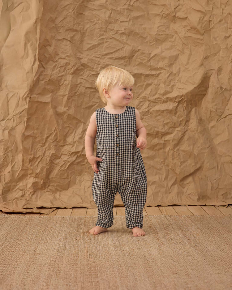 A toddler with blond hair models the rylee + cru button jumpsuit in black check, barefoot on a woven rug, one hand in their pocket and smiling slightly against a crumpled brown paper backdrop.