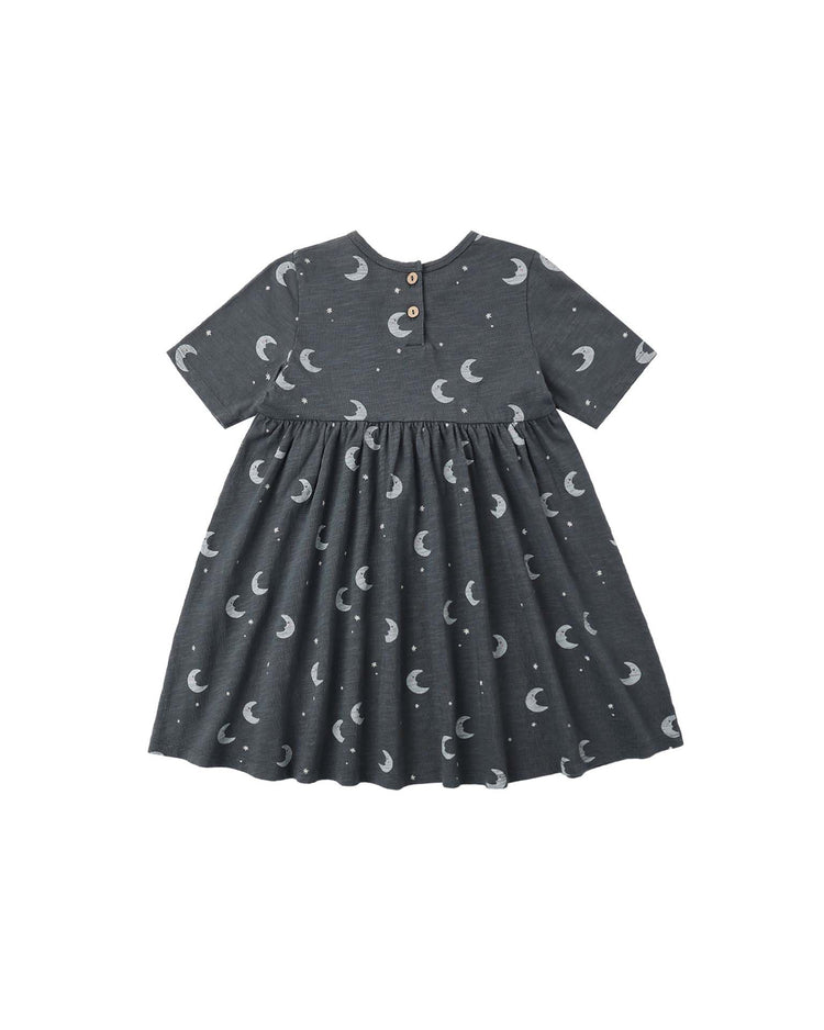 The PREORDER finn dress in moons by rylee + cru is a dark gray, short-sleeve dress for children, made from soft cotton jersey. It features a gathered skirt adorned with an allover moon and dot pattern and has two small buttons at the back neckline. The dress is shown on a plain white background.
