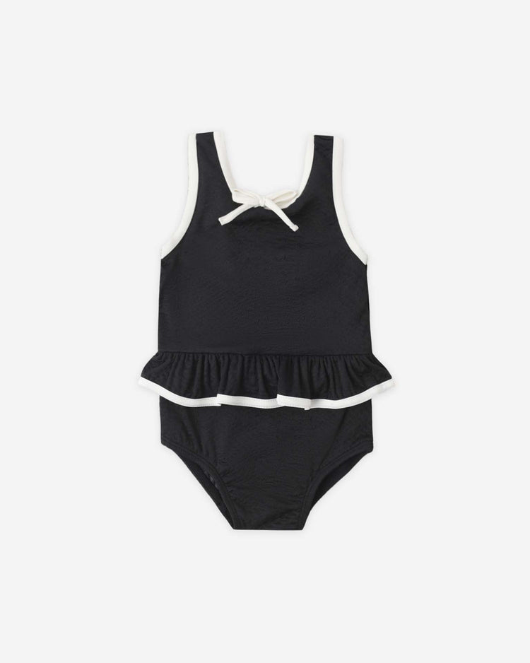 The rylee + cru skirted one-piece in black is a sleeveless toddler swimsuit with white trim, a bow at the neckline, a ruffled waist skirt, and UPF 50+ sun protection.