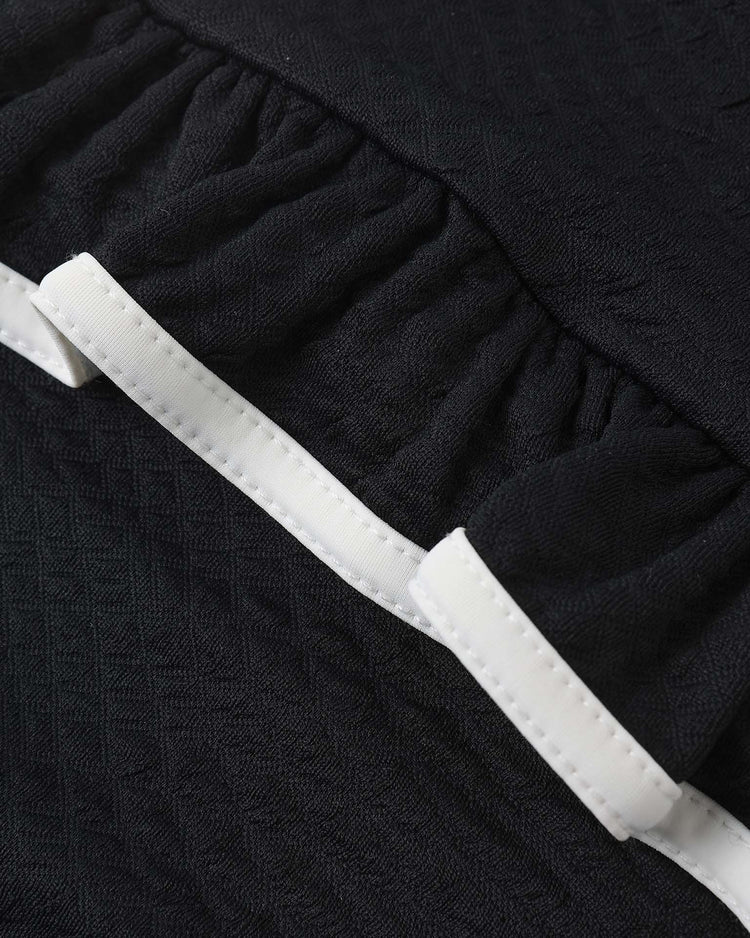 Close-up of rylee + cru's skirted one-piece in black, featuring textured fabric with gathered ruching and a white ribbon near the edge; offers UPF 50+ sun protection.