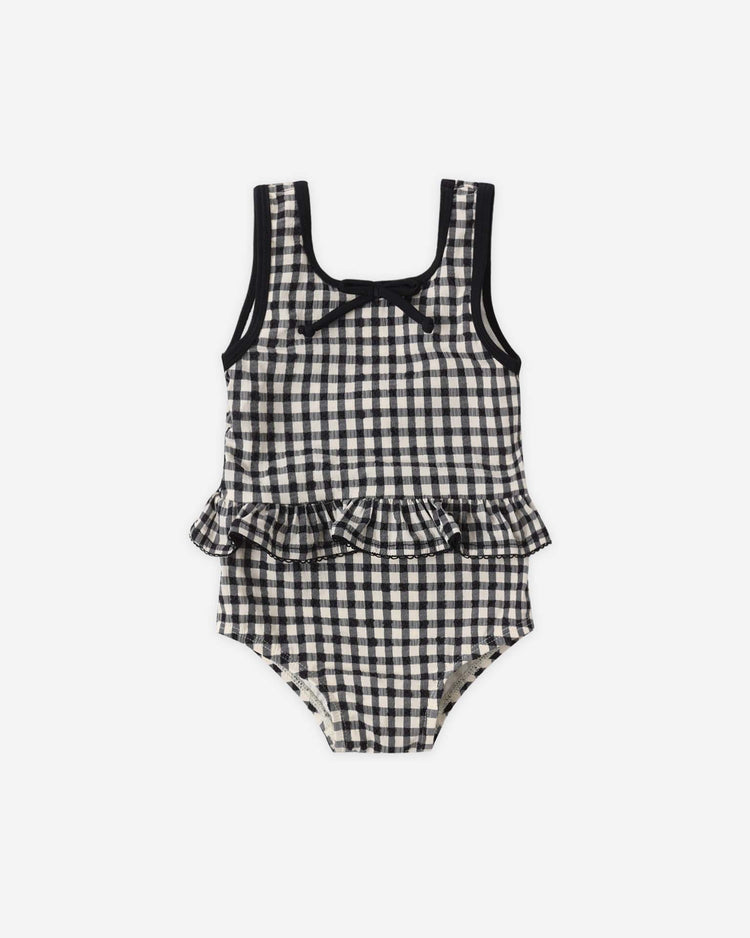 The rylee + cru skirted one-piece in black gingham features black trim, a chest bow, peplum ruffle at the waist, and built-in UPF 50+ sun protection.