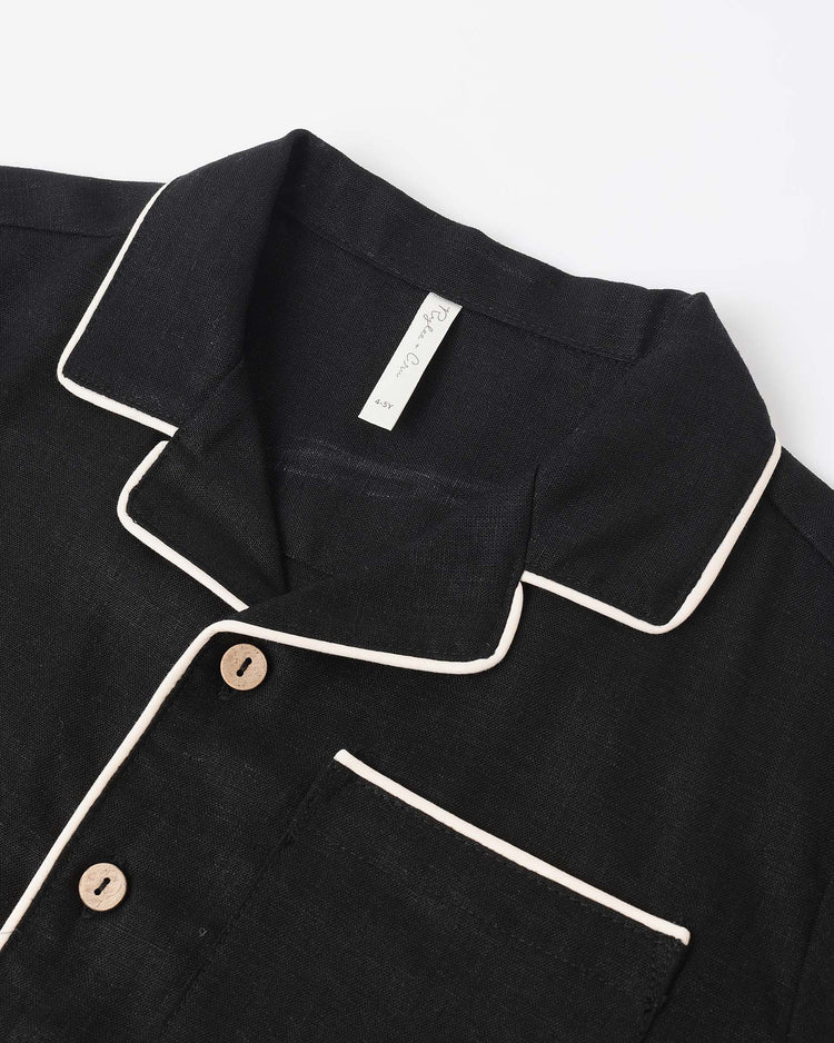 Close-up of rylee + cru's lapel collared shirt in black featuring white piping on the collar, lapel, and pocket, light brown buttons, and a visible white tag inside the collar.