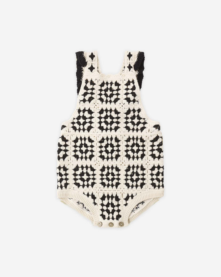 The rylee + cru crochet romper in abstract is a sleeveless 100% cotton piece, featuring black and white square motifs, button closures at the bottom, and black crochet shoulder details, shown on a white background.