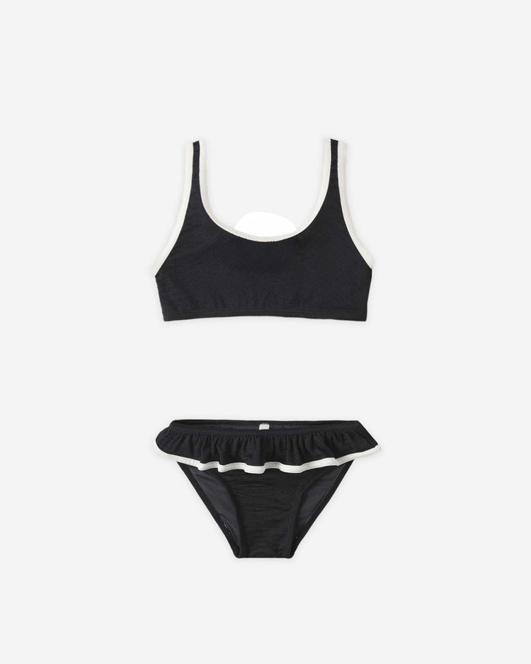 The daisy bikini in black by rylee + cru is a two-piece set with a scoop neck top, high-cut bottoms, white trim, ruffled waistband, UPF 50+ protection, and is made from recycled polyamide. Shown on a plain white background.