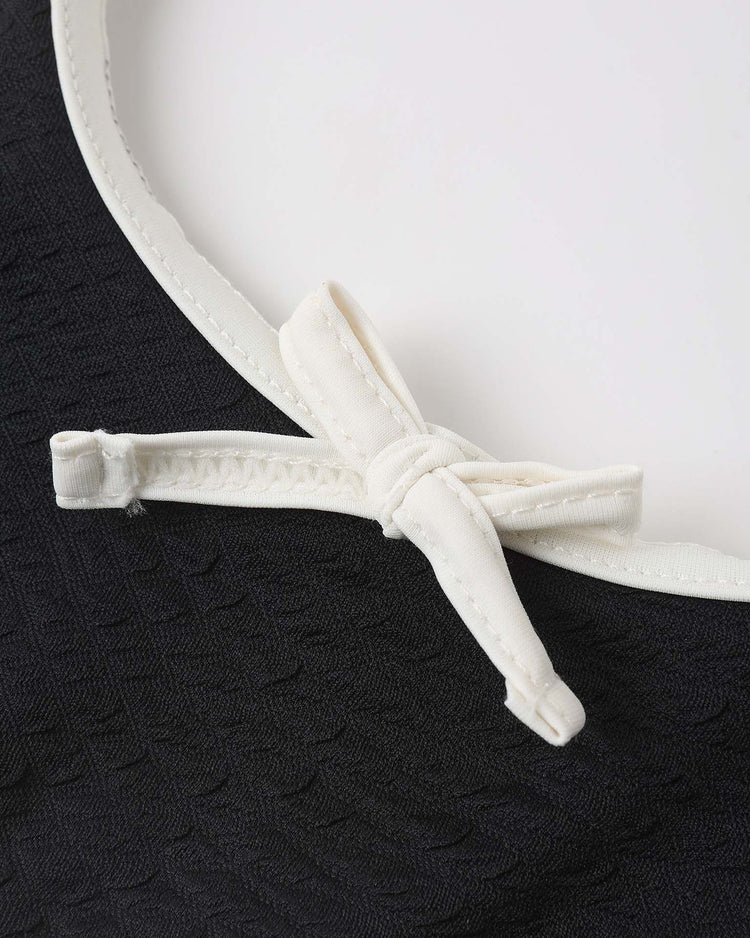 Close-up of the daisy bikini in black by rylee + cru, showing its textured recycled polyamide fabric with a white border and a small white bow on the edge, set against a plain white background.