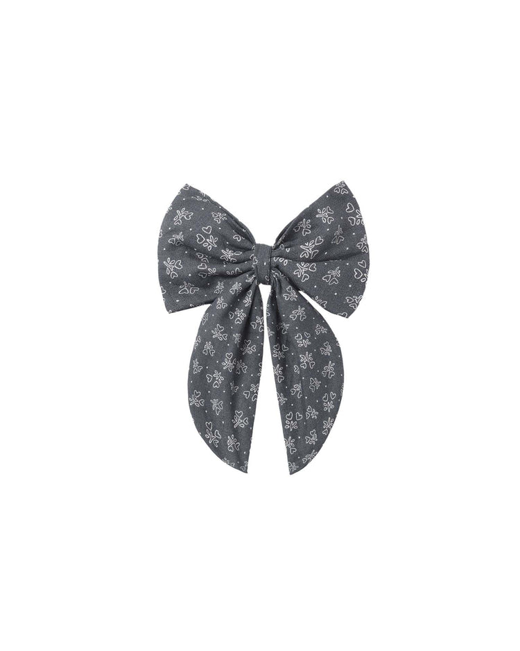 The rylee + cru oversized bow in heart ditsy is a dark gray hair accessory adorned with a subtle white heart ditsy print. Featuring two large loops and two long, wide tails, this elegant bow is attached to an alligator clip and displayed against a plain white background.