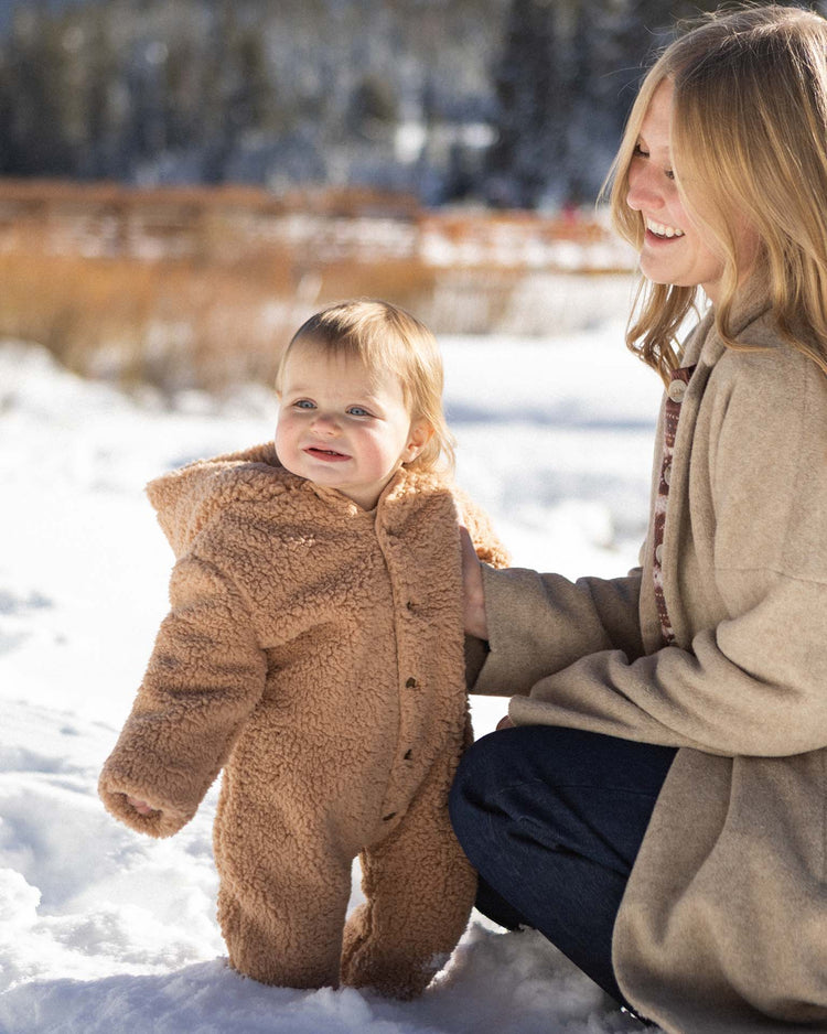 A smiling woman kneels in the snow beside a baby bundled up in the rylee + cru fleece jumpsuit in fawn. Sunlight streams through trees and a wooden fence in the background, highlighting the cozy moment.